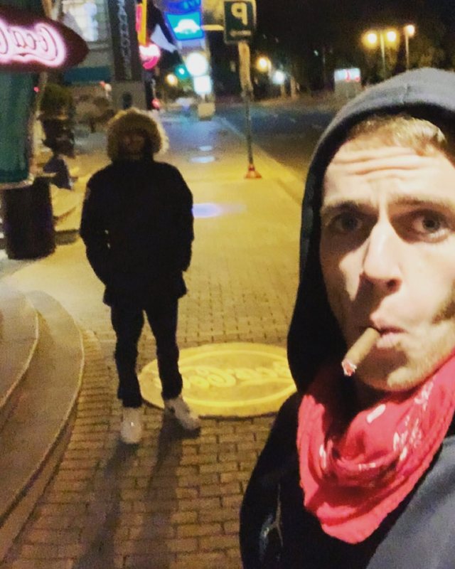 Alex on a city street at night in a dark hoodie, smoking, with a hooded figure standing behind him