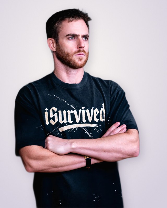 Alex today, arms crossed, wearing a shirt that reads iSurvived