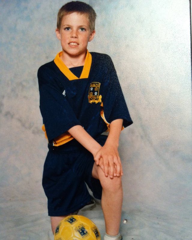 Alex as a boy in his soccer uniform, kneeling beside a yellow soccer ball